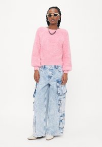 Fluffy roze sweater met lange mouwen, gecombineerd met brede, lichtblauwe cargobroeken met zakken en ritsen. Model draagt witte schoenen.