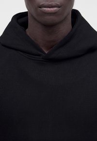 Gros plan d'une personne portant un sweat à capuche noir uni, montrant la partie inférieure du visage, le cou et le haut de la poitrine.