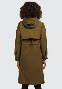 Giacca impermeabile lunga di colore marrone con vita cinchata, cappuccio oversize e patch con logo nero. Presenta maniche con bottoni e una texture liscia.