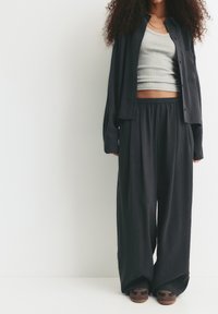 Person wearing loose black pants, black button-up shirt over a gray crop top, and brown sandals, standing against a plain white background.