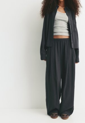 Person wearing loose black pants, black button-up shirt over a gray crop top, and brown sandals, standing against a plain white background.