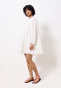 Langes, weißes, fließendes Kleid mit gerafftem Oberteil und langen Ärmeln, kombiniert mit roten flachen Sandalen mit gekreuzten Riemen. Glatter, leichter Stoff.