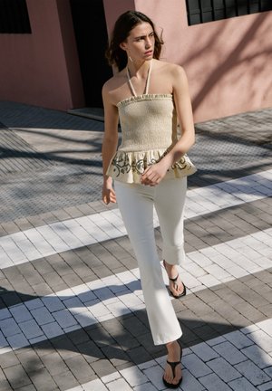 Frau, die draußen einen beigen gerafften Neckholder-Top mit gemustertem Peplum, weiße Hosen und schwarze Sandalen im Sonnenlicht und Schatten trägt.