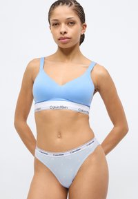 Απαλό γαλάζιο μπουστάκι με V-λαιμόκοψη και λεπτές τιράντες, διακοσμημένο με λευκή ταινία λογότυπου Calvin Klein. Συνδυάζεται με γαλάζι ο σλιπ μπραζίλιαν με σχέδια.