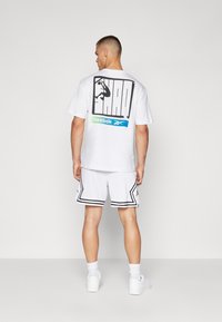 Hvid kortærmet skjorte med et stort sort Shaq-logo på ryggen, kombineret med hvide mesh-shorts med sorte detaljer og kant.