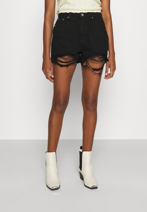 Personne portant un short en jean noir déchiré, un haut sans manches couleur claire et des bottines blanches avec des côtés élastiques noirs.