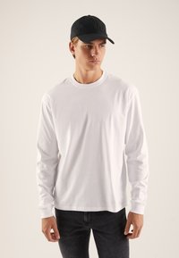 Hvid langærmet t-shirt, lavet af bomuld, med rund hals; kombineret med en sort baseballkasket og mørke jeans. Enkel design, ingen grafik.