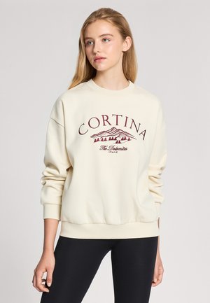 Maglione crema con collo rotondo, polsini e orlo a costine. Presenta una grafica color bordeaux di montagne e il testo "CORTINA". Texture morbida.