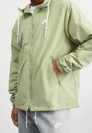 Personne portant une veste zippée Nike vert clair avec capuche et un pantalon de survêtement Nike gris avec cordons blancs apparents.