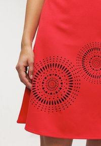 Rotes Kleid aus glattem Stoff mit lasergravierten runden Mustern in schwarzer Detailierung, mit einer A-Linie Silhouette und sichtbarem Saum.