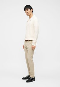 Άνδρας μοντέλο στέκεται, φορώντας εκρού πλεκτό πουλόβερ, μπεζ παντελόνι, καφέ ζώνη και μαύρα δερμάτινα loafers, σε απλό λευκό φόντο.