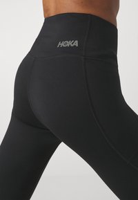 Hochtaillierte schwarze Leggings aus dehnbarem Stoff, mit Flachnaht und einem HOKA-Logo am Bund. Geschmeidige Textur.