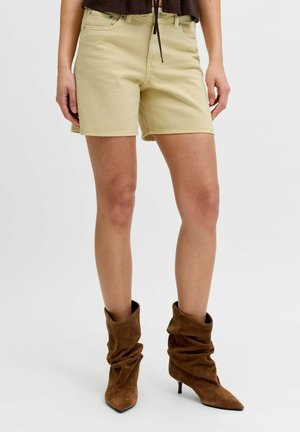 Nő bézs színű farmer rövidnadrágban, barna laza, hegyes orrú, közepes sarkú csizmával, egyszínű háttér előtt.