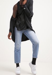 Osoba ve svetle modrých džínách, černém topu, černém cardiganovém svetru spadlém z jednoho ramene, černých ponožkách a bílých teniskách, stojící s jednou nohou zvednutou.