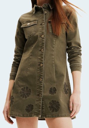 Kobieta ubrana w oliwkową sukienkę na guziki z kieszeniami na piersiach i haftowanymi kwiatowymi wzorami w pobliżu brzegu, rękawy podciągnięte.