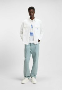 Hvid knap-op jakke med to brystlommer, parret med en lyseblå grafisk t-shirt og lyseblå jeans. Fodtøjet er hvide sneakers.