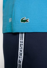 Turkos kortärmad skjorta med "ULTRA DRY" etikett och grön broderad krokodil-logotyp, matchad med marinblå byxor med vit "LACOSTE" rand.