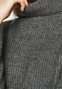 Suéter gris de punto con un cuello alto y enrollado. La textura de la tela es acanalada, con una mezcla de hilos claros y oscuros que le dan profundidad.