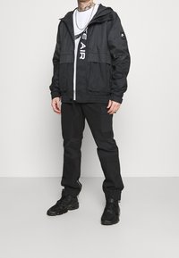 Svart vindjacka med huva, dragkedja framtill och elastiska ärmslut. I kombination med svarta cargobyxor och sneakers, med en logo på bröstet.