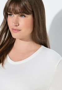 Bílý bavlněný top s výstřihem do V, hladkým povrchem, jemným zakončením a decentním švem. Modelka s dlouhými hnědými vlasy a lehkým líčením.