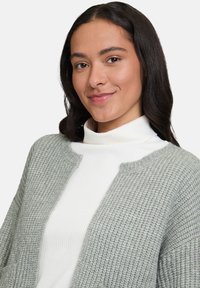 Jeune femme aux cheveux bruns portant un col roulé blanc côtelé et un cardigan texturé gris, souriant légèrement devant un fond uni.