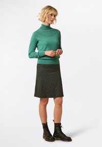 Grüner Rollkragenpullover mit strukturiertem Detail am Kragen, kombiniert mit einem gemusterten knielangen Rock, der geometrische Formen in Orange und Türkis zeigt. Klobige schwarze Schnürstiefel runden das Outfit ab.