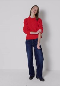 Roter gestrickter Pullover mit Rundhalsausschnitt, kombiniert mit dunkelblauen weit geschnittenen Jeans. Das Model steht mit verschränkten Armen vor einem neutralen Hintergrund.