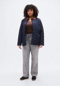 Σκουρό μπλε μπουφάν με επένδυση καπιτονέ και λεπτομέρεια καφέ γιακά, με κουμπιά snap και τσεπάκι με φερμουάρ, σε συνδυασμό με ανοιχτό γκρι τζιν και μαύρα παπούτσια.