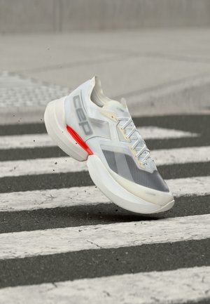 White and gray athletic sneaker with red accent and CAP logo, mid-air above a pedestrian crosswalk on asphalt pavement.