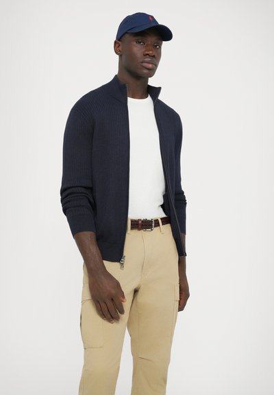 Cardigã azul-marinho com fecho de correr e malha canelada, camisa branca de gola redonda, calças cargo cor de caramelo e cinto castanho entrelaçado, usados com um boné azul-marinho com um logótipo.