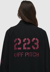 Svart tröja med en texturerad yta. Har stora vinröda siffror "223" och "OFF PITCH" i fet stil på ryggen.