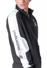 Chaqueta negra con una franja blanca en una manga que presenta el texto "Motorsport". Con cremallera en la parte frontal, tejido suave, diseño deportivo.