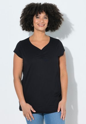 Lächelnde Frau mit lockigem Haar, die ein schwarzes kurzärmeliges Oberteil und blaue Jeans trägt, vor einem schlichten weißen Hintergrund stehend.