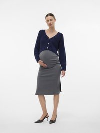 Gravid kvinna i en marinblå kofta och grå pennkjol, som står och försiktigt håller om sin mage, iklädd svarta kitten heels.