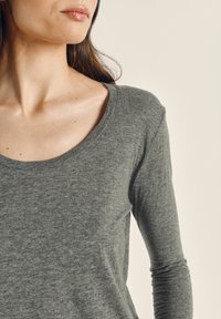 Moteris su ilgais rudais plaukais, dėvinti pilką ilgomis rankovėmis palaidinę su apvalia iškirpte, šiek tiek pasukta į kairę, ant smėlio spalvos fono.