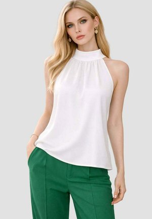 Femme blonde portant un haut blanc sans manches à col montant et un pantalon vert, debout avec une main dans la poche devant un fond uni.