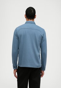 Modrá bavlněná košile s dlouhými rukávy, zapínacími manžetami a rovný střih. Obsahuje ležérní límec a viditelné švy na zadní straně.