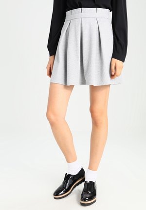Femme portant une jupe plissée gris clair taille haute, un haut noir à manches longues, des chaussettes blanches et des chaussures plateformes noires brillantes.