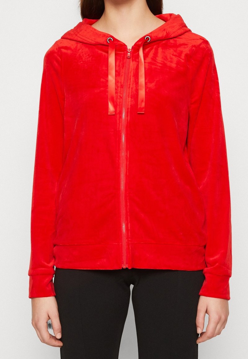 Sudadera roja con cremallera, confeccionada en una tela suave y texturizada, con capucha, cordones de satén y bolsillos laterales. Cuenta con mangas largas y un ajuste holgado.
