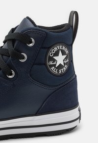 Vysoký teniskový model v námořnické modři s texturovanou kůží a látkou. Má černou kruhovou nášivku s logem a bílou gumovou podrážku s černými prvky.