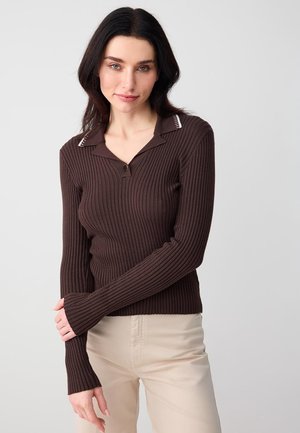 Giovane donna dai capelli scuri che indossa un top marrone a coste con maniche lunghe e pantaloni beige chiaro, in piedi contro uno sfondo grigio uniforme.