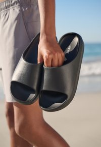 Schwarze Slip-On-Sandalen mit offenem Zeh-Design, aus strukturiertem Material, in der Hand gehalten vor einem Strandhintergrund, zeigen Details der Innensohle.