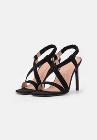 Black suede high-heeled sandals with crisscross straps and a cushioned insole. Features a slender heel and a soft elasticized back strap.