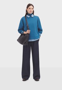 Felpa blu oversized indossata sopra una camicia colletto azzurra, abbinata a pantaloni in denim scuri a gamba larga e una borsa a tracolla nera.