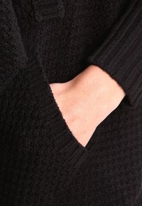 Ruka vložená do přední kapsy černého pleteného svetru s texturou a žebrovaným manžetovým lemem na rukávu.