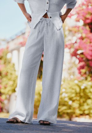 Personne portant un pantalon large gris clair avec taille à cordon de serrage et une chemise boutonnée à manches courtes assortie, debout à l'extérieur sur une surface pavée.