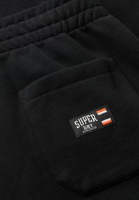 Zwarte katoenen shorts met een achterzak en een rechthoekig logolabel met de tekst "SUPER DRY ATHLETICS" in wit, oranje en zwart.