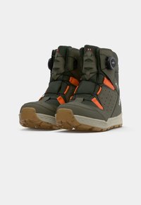 Une paire de bottes de randonnée vert olive et orange avec des semelles beiges et des dispositifs de laçage à molette, prises sous un angle pour montrer le devant et les côtés extérieurs sur fond blanc.