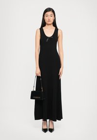 Woman wearing a sleeveless black maxi dress with a small bow, holding a black "Love Moschino" handbag, and wearing black pointed heels.