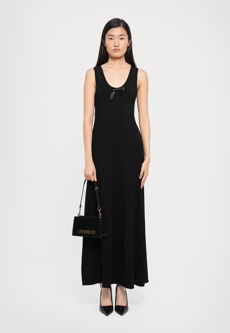 Woman wearing a sleeveless black maxi dress with a small bow, holding a black "Love Moschino" handbag, and wearing black pointed heels.